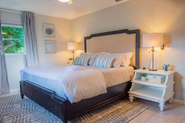 A bedroom with a dark wooden bed, white bedding, two bedside lamps, a white nightstand, and a view of greenery outside.