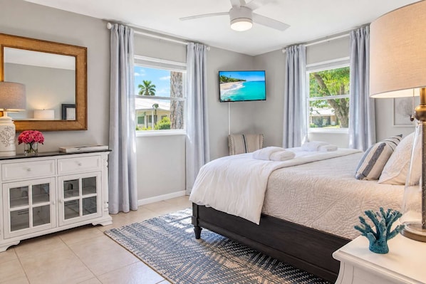 A bedroom with a bed, a TV, a ceiling fan, and a view of the outdoors.