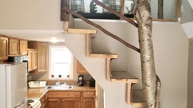 Una cocina de varios niveles con armarios de madera, un refrigerador y una estufa. Una escalera de madera con diseño de tronco de árbol conduce a un nivel superior.