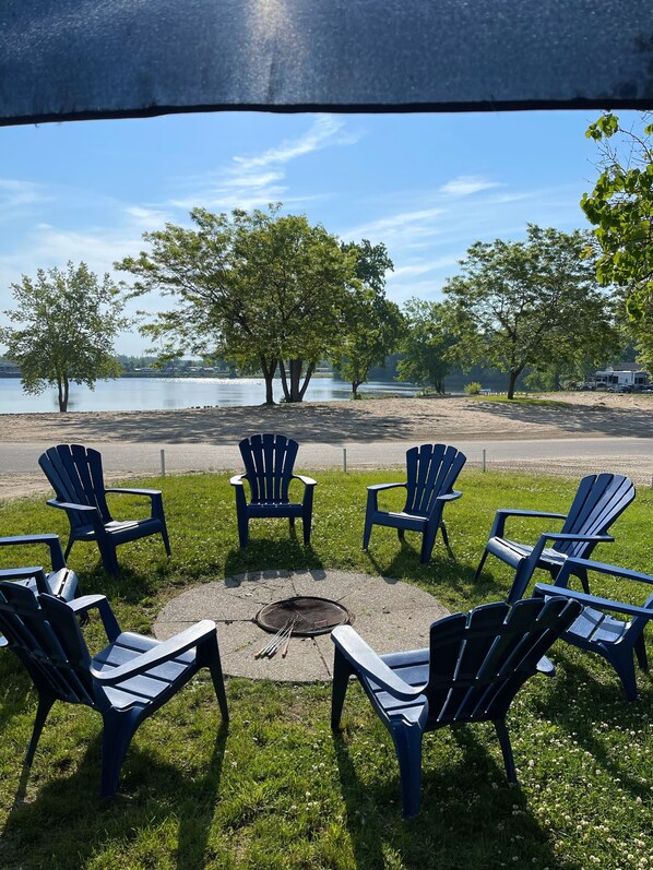 A lakeside seating area with Adirondack chairs arranged around a fire pit, overlooking a calm lake and trees. - Waterfront at Sherkston Shores Resort (Port Colborne)