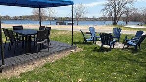 An outdoor patio with a table and chairs under a blue canopy, overlooking a lake with a dock and picnic table in the background.