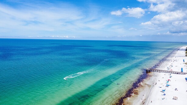 Vista aérea de una playa con aguas turquesas cristalinas, una orilla arenosa y un muelle que se adentra en el agua.