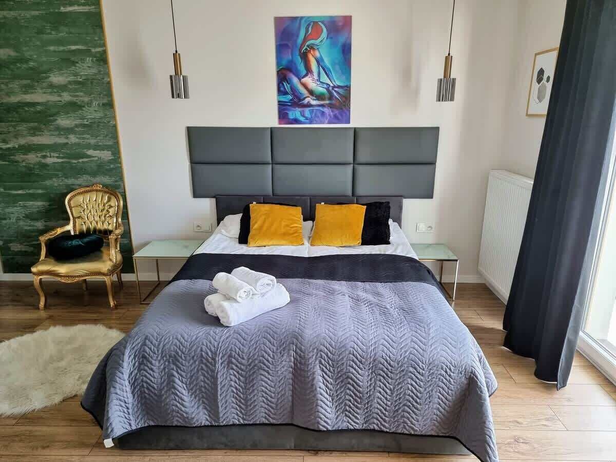 Una cama bien hecha con colcha gris, dos almohadas y un juego de toallas. Hay un cuadro en la pared sobre la cama, una silla y una ventana con cortinas.
