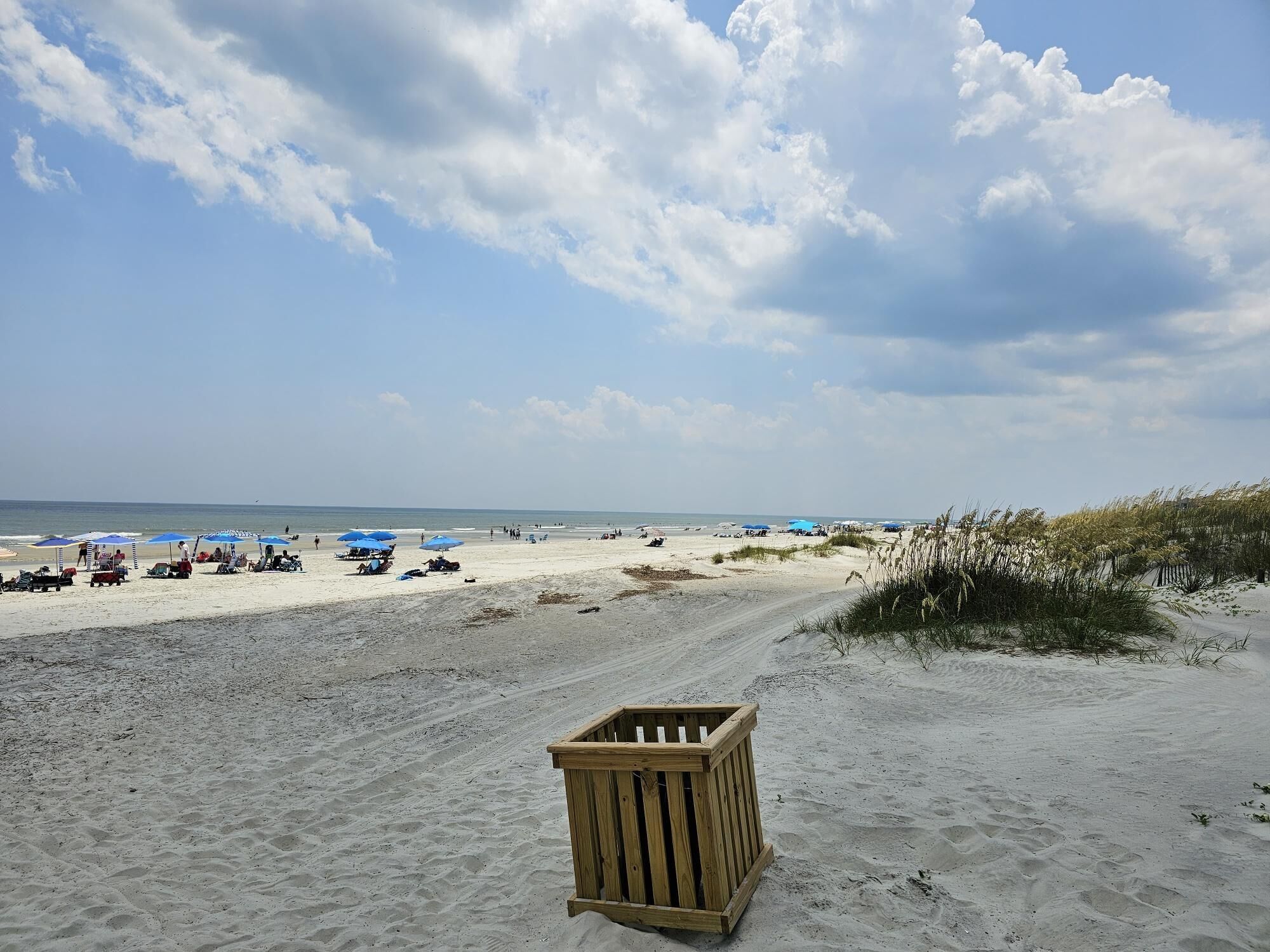 A sandy beach with a wooden structure in the foreground, a clear sky, and beach umbrellas in the distance.
