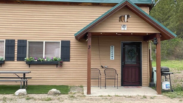 Une cabane avec un toit végétalisé, un revêtement extérieur en bois, une pancarte de bienvenue et un banc à l’extérieur.