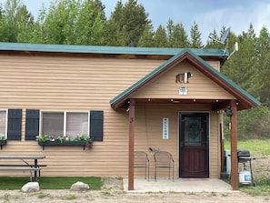 A cabin with a green roof, a wooden exterior, a welcome sign, and a bench outside.