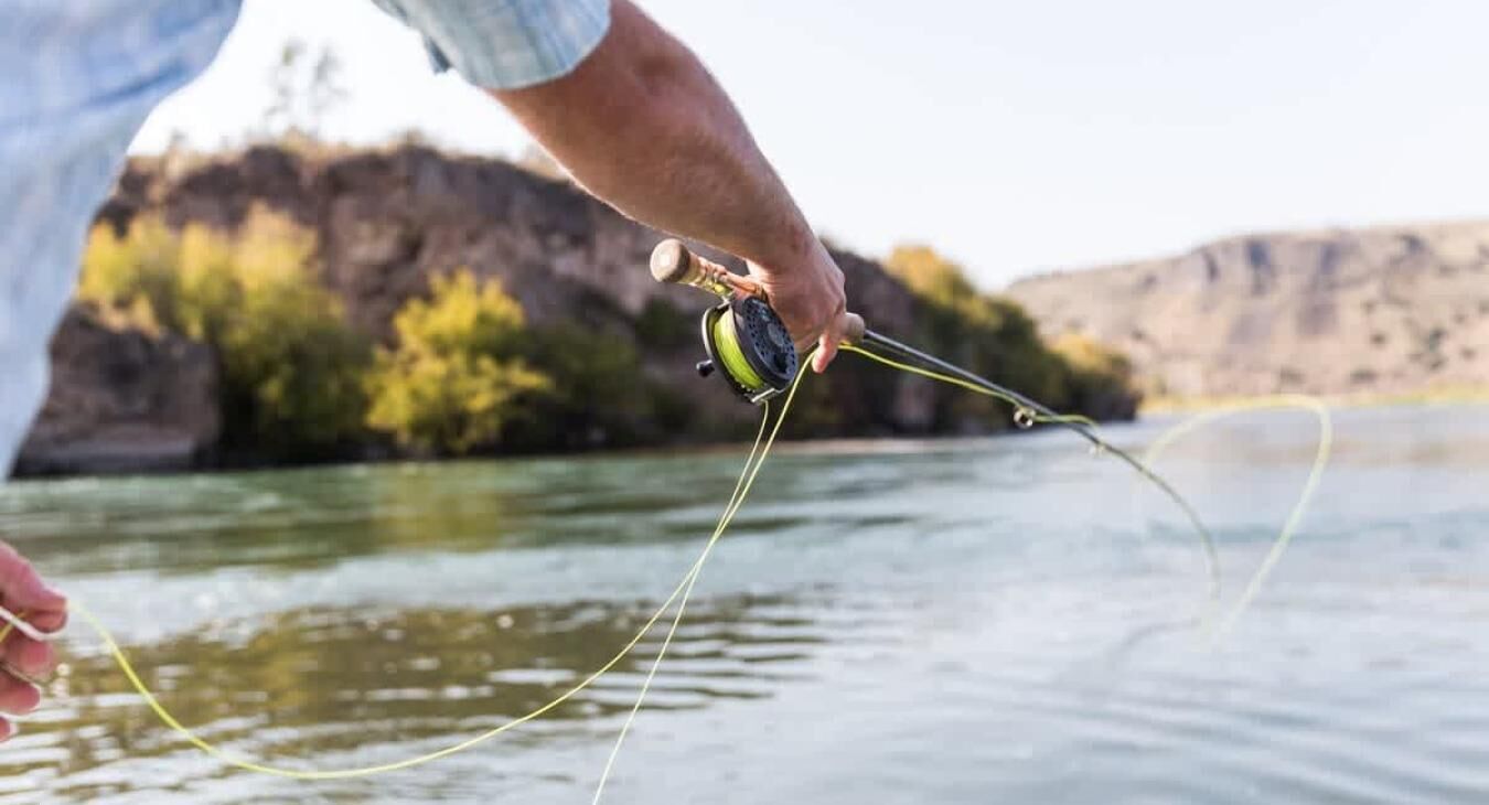A person holding a fishing rod with a reel, with a body of water and a rocky landscape in the background.