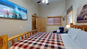 Une chambre avec une couvre-lit à carreaux, une tête de lit en bois, une fenêtre avec des stores, un tableau représentant un ours et une porte donnant sur une autre pièce.