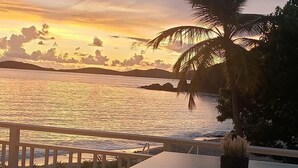 A wooden deck with a table and two stools overlooking a body of water at sunset. - Darling Palms Tree House: Beachside Studio Oasis (St Thomas)
