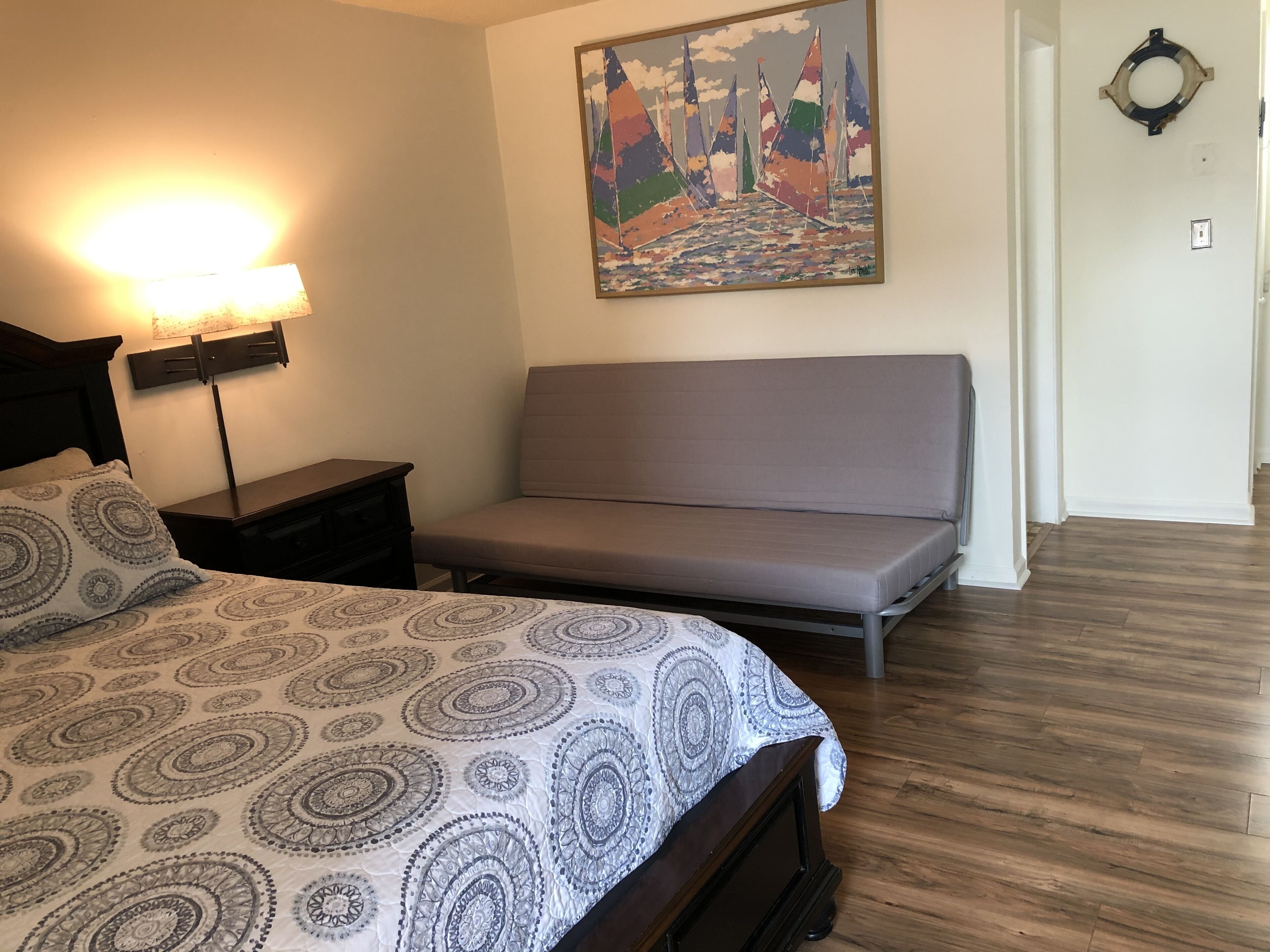 A bedroom with a bed, a bedside table, a lamp, a gray sofa, and a painting on the wall.