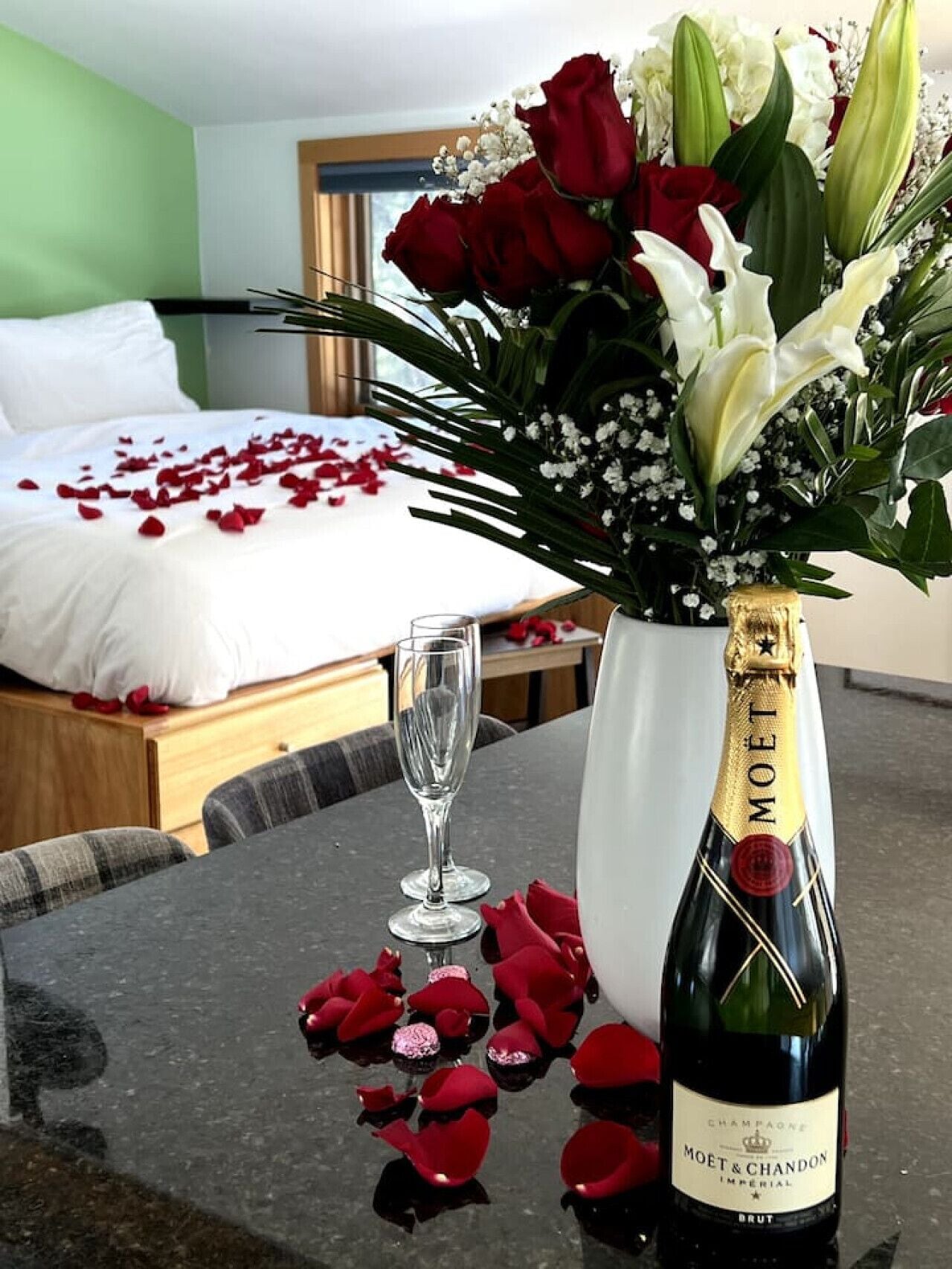 Una habitación de hotel con cama, un jarrón de flores, una botella de champán y pétalos de rosa en el suelo.