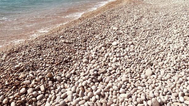 A pebbled beach with clear blue water and a rocky cliff in the background.