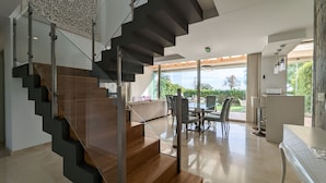 A modern interior with a wooden staircase, a dining area with chairs and a table, and a view of greenery outside.