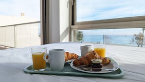 A breakfast tray with croissants, orange juice, and coffee on a table by a window overlooking the sea.