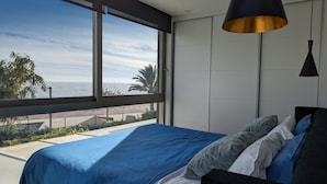 A modern bedroom with a large window overlooking a beach, a bed with blue bedding, and a black pendant light.