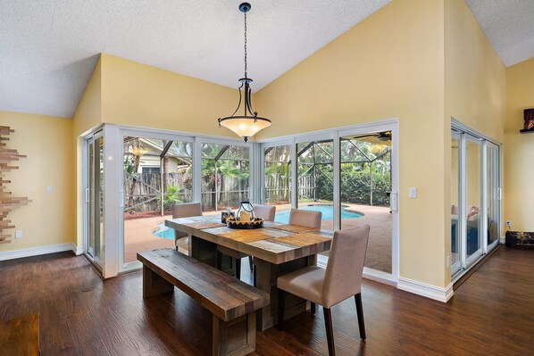 Enjoy meals in this dining area with a large table for four and a bench, surrounded by a high ceiling and large glass doors leading to an outdoor space.