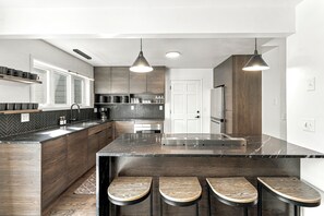 A modern kitchen with a large island, bar stools, pendant lighting, and a herringbone backsplash.