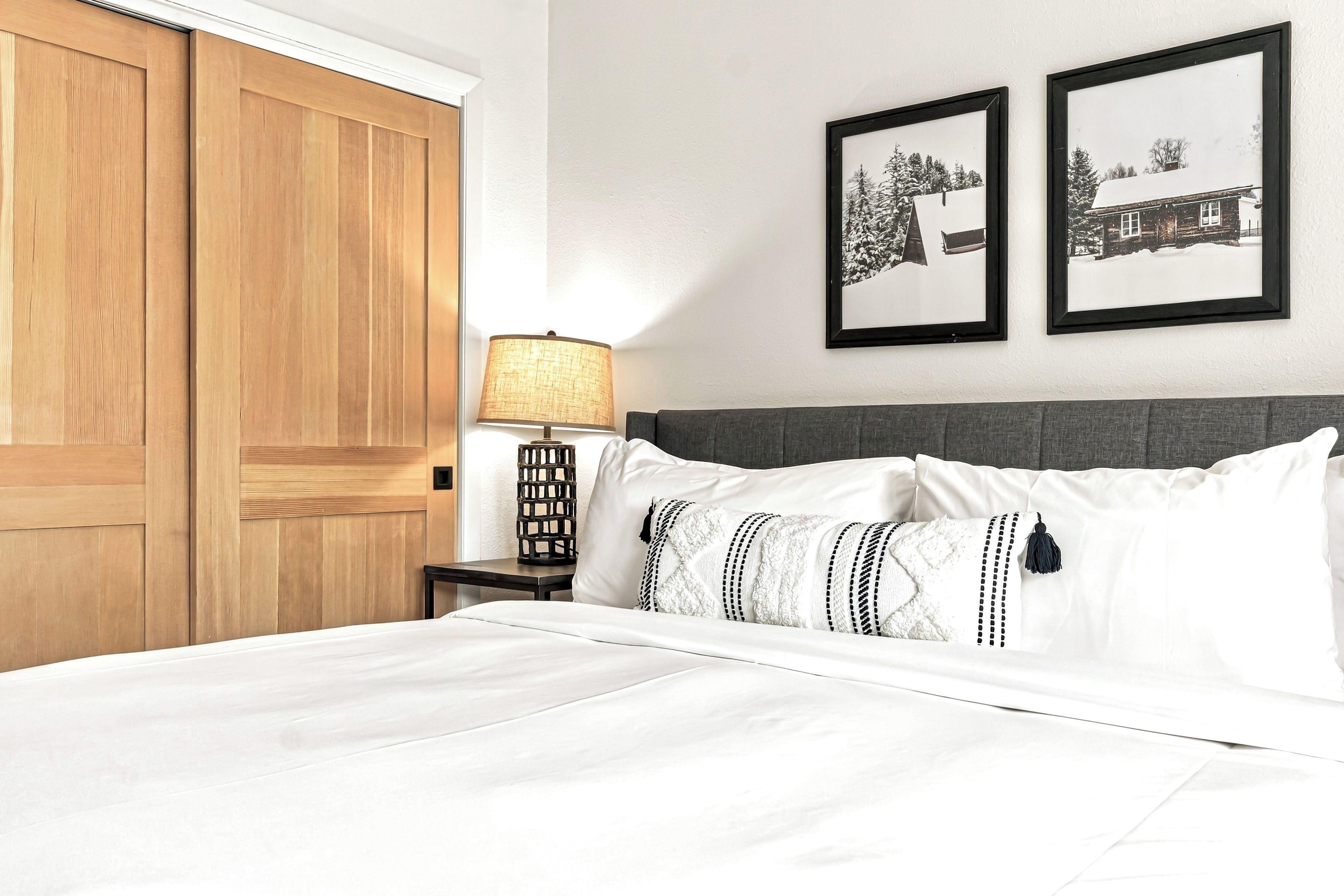 A hotel room with a large wooden wardrobe, a bedside table with a lamp, a bed with white bedding, and two framed pictures on the wall.