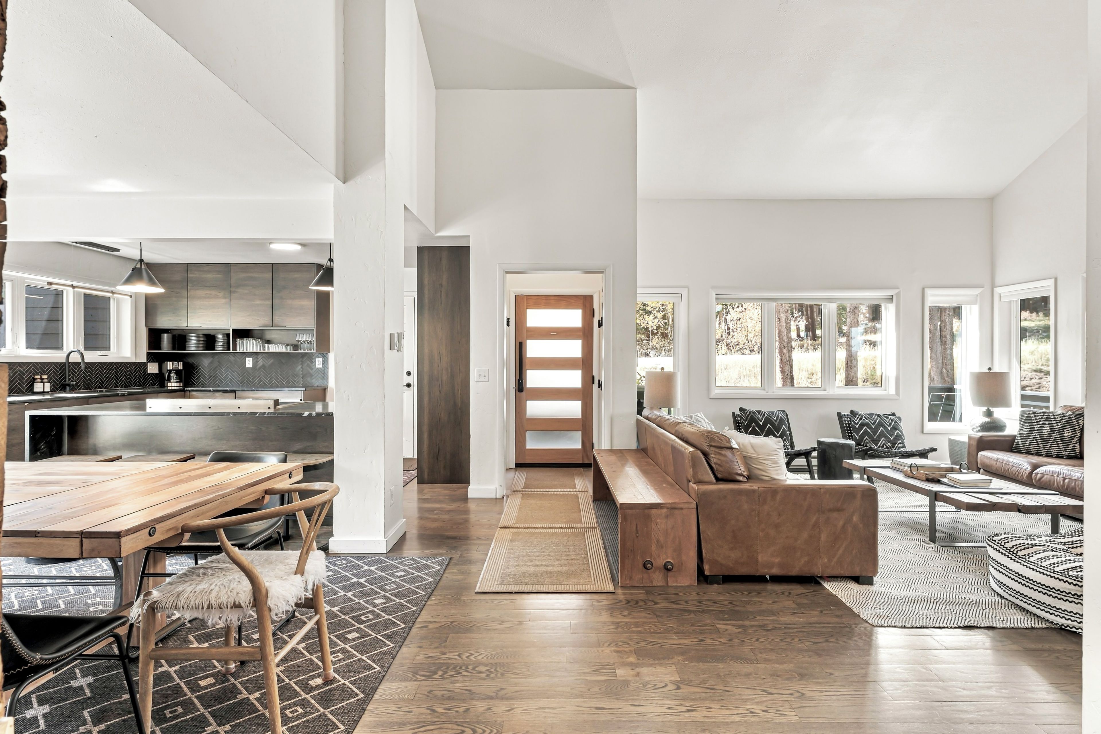 A spacious living area with a wooden dining table, a sofa, and a coffee table. There is a kitchen area with modern appliances and a large window.