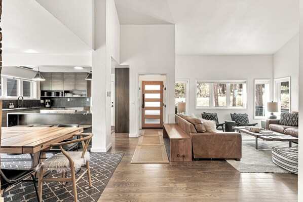 A spacious living area with a wooden dining table, a sofa, and a coffee table. There is a kitchen area with modern appliances and a large window.