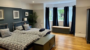 Un dormitorio con cama, mesita de noche, banqueta y ventana con cortinas.