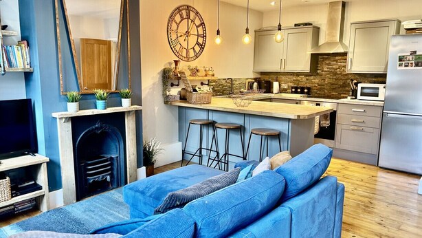 Una sala de estar moderna con un sofá azul, una chimenea y una zona de cocina con un reloj grande y lámparas colgantes.