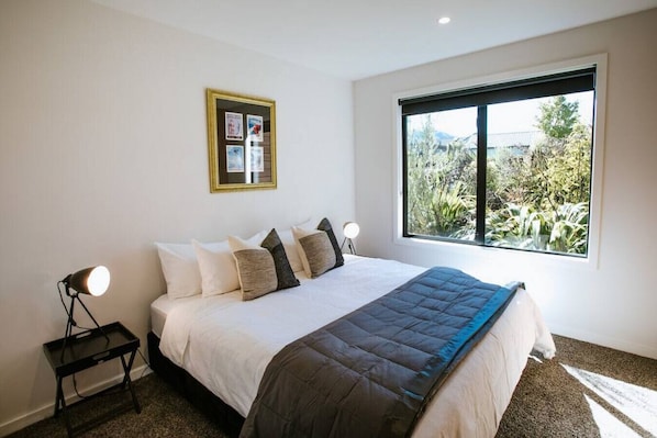 A bedroom with a large bed, a window with a view of greenery, a bedside table with a lamp, and a framed picture on the wall.