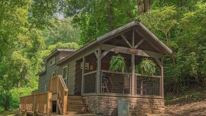 Une cabane rustique avec des fondations en pierre, une terrasse en bois et un porche couvert agrémenté de plantes suspendues.