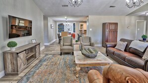 A spacious living room with a large sofa, a coffee table, and a fireplace. There is a dining area in the background with a chandelier.