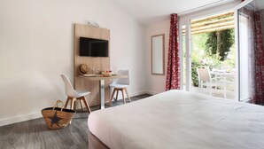 A hotel room with a bed, a desk with a TV, two chairs, a basket, and a view of the garden through the window. - Studio Rustic | Accès Piscine Saisonnière (Arles)