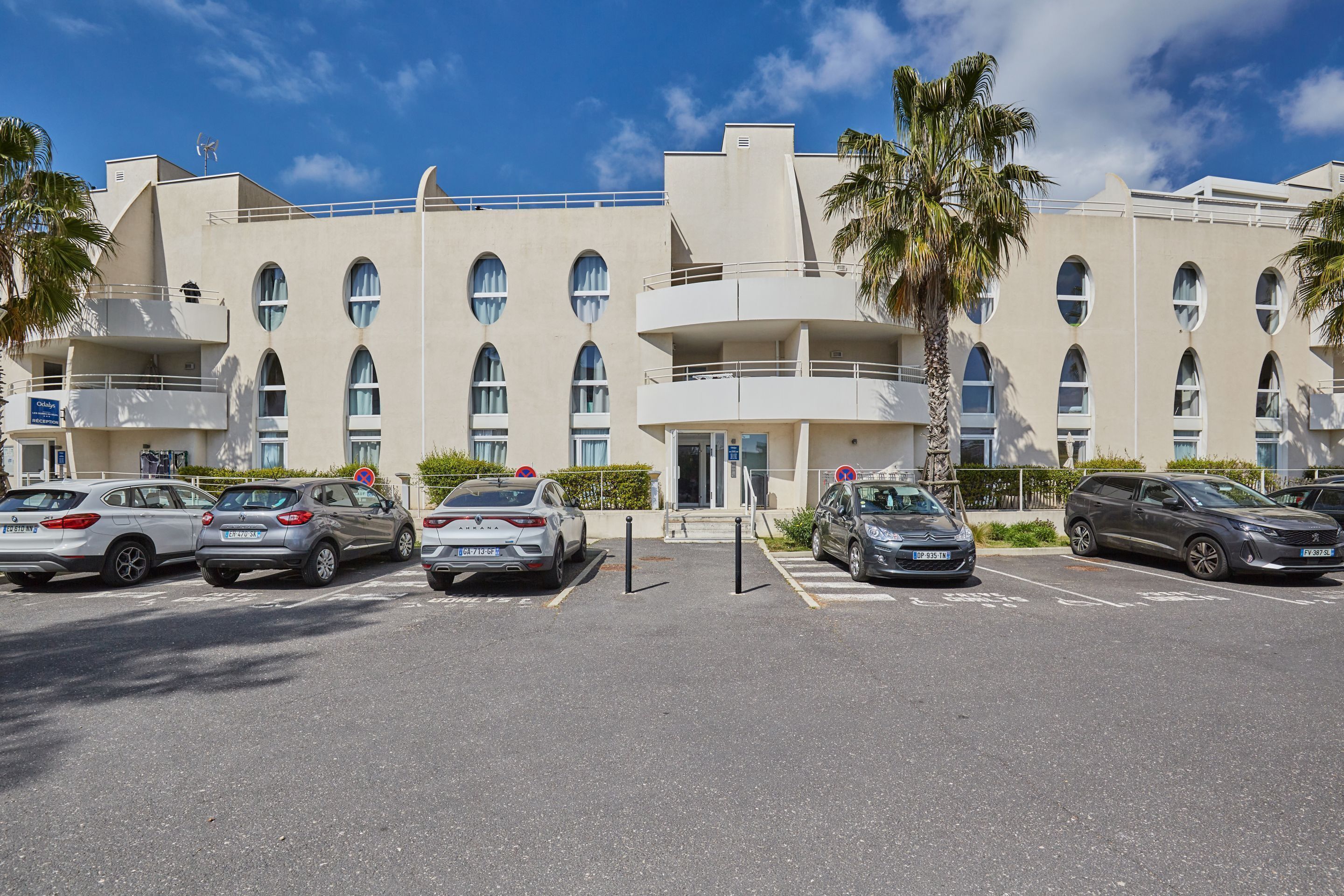 A modern hotel with a parking lot, multiple cars, and a clear blue sky.