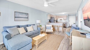 Een moderne woonkamer met een bank, salontafel en stoelen, een keuken met een centraal kookeiland, en een plafondventilator.