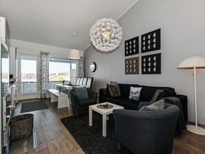 Uma sala de estar moderna com sofá, poltrona, mesa de centro e área de jantar com mesa e cadeiras. Há uma janela grande com cortinas, luminária no teto e obras de arte nas paredes.