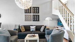 A modern living room with a grey sofa, two armchairs, a white coffee table, and a chandelier. There is a staircase with a wooden railing leading to an upper level. - Skivillas 78 ukkohalla (2) by Interhome (Hyrynsalmi)