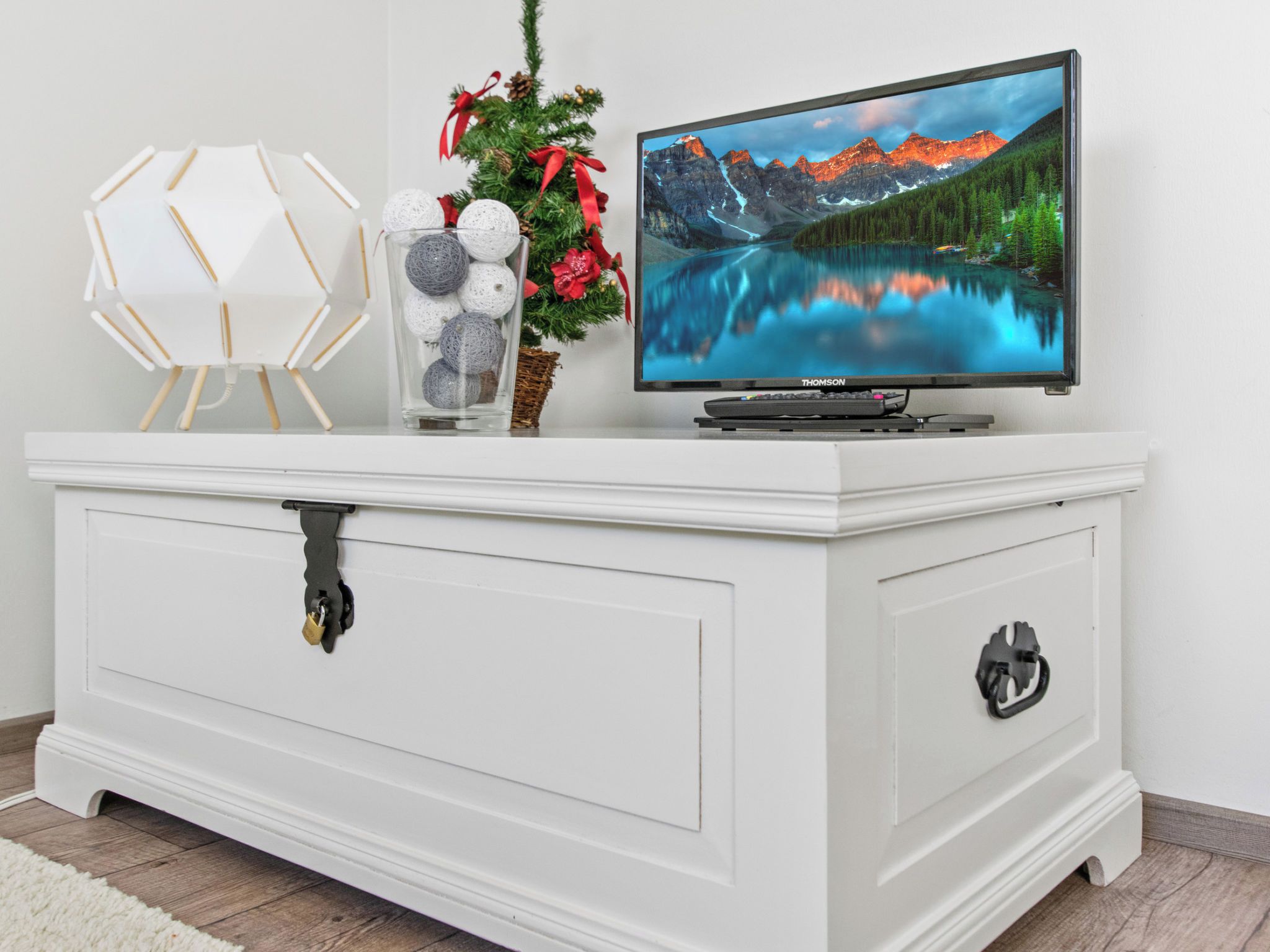 Un meuble de rangement blanc pour appareils électroniques, avec un téléviseur à écran plat affichant un paysage de montagne, un vase décoratif en verre orné de boules, et un petit sapin de Noël décoré de rubans rouges.