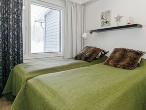 Un dormitorio con dos camas, una ventana con persianas, un estante montado en la pared con objetos decorativos y una lámpara.