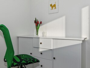 Una habitación con un escritorio blanco, una silla verde, un jarrón con flores rojas y un cuadro con la imagen de un gato.