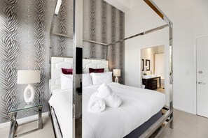 Moderni hotellihuone, jossa on nelipylväinen sänky, suuri peili ja kylpyhuone, joka näkyy avoimesta ovesta.