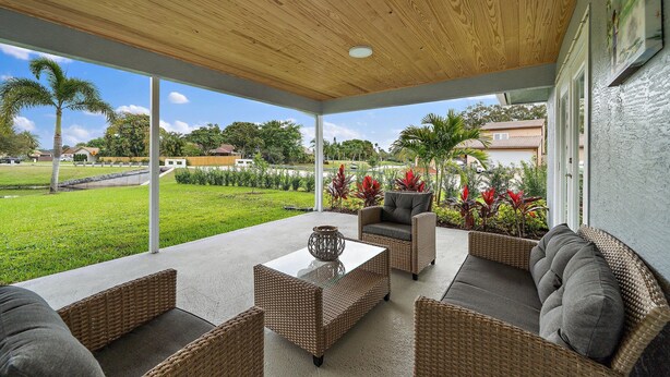 Een overdekt terras met rieten meubels en een betonnen tafel, met uitzicht op een grasachtig landschap met palmbomen en huizen.
