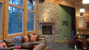 Un salón acogedor con chimenea de piedra, techo de madera y ventanas amplias con vistas al bosque.