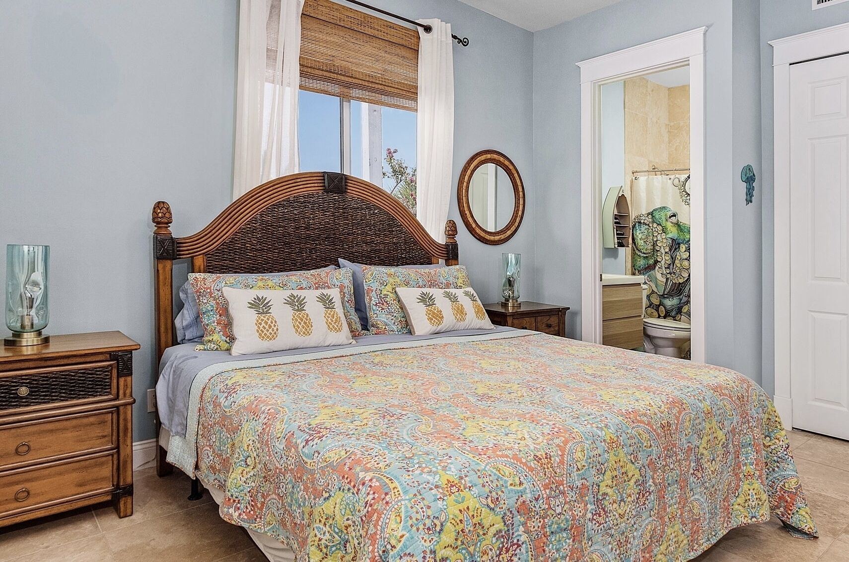 Une chambre avec un grand lit, une table de chevet en bois, une commode, un miroir rond et une fenêtre avec un store en bambou.