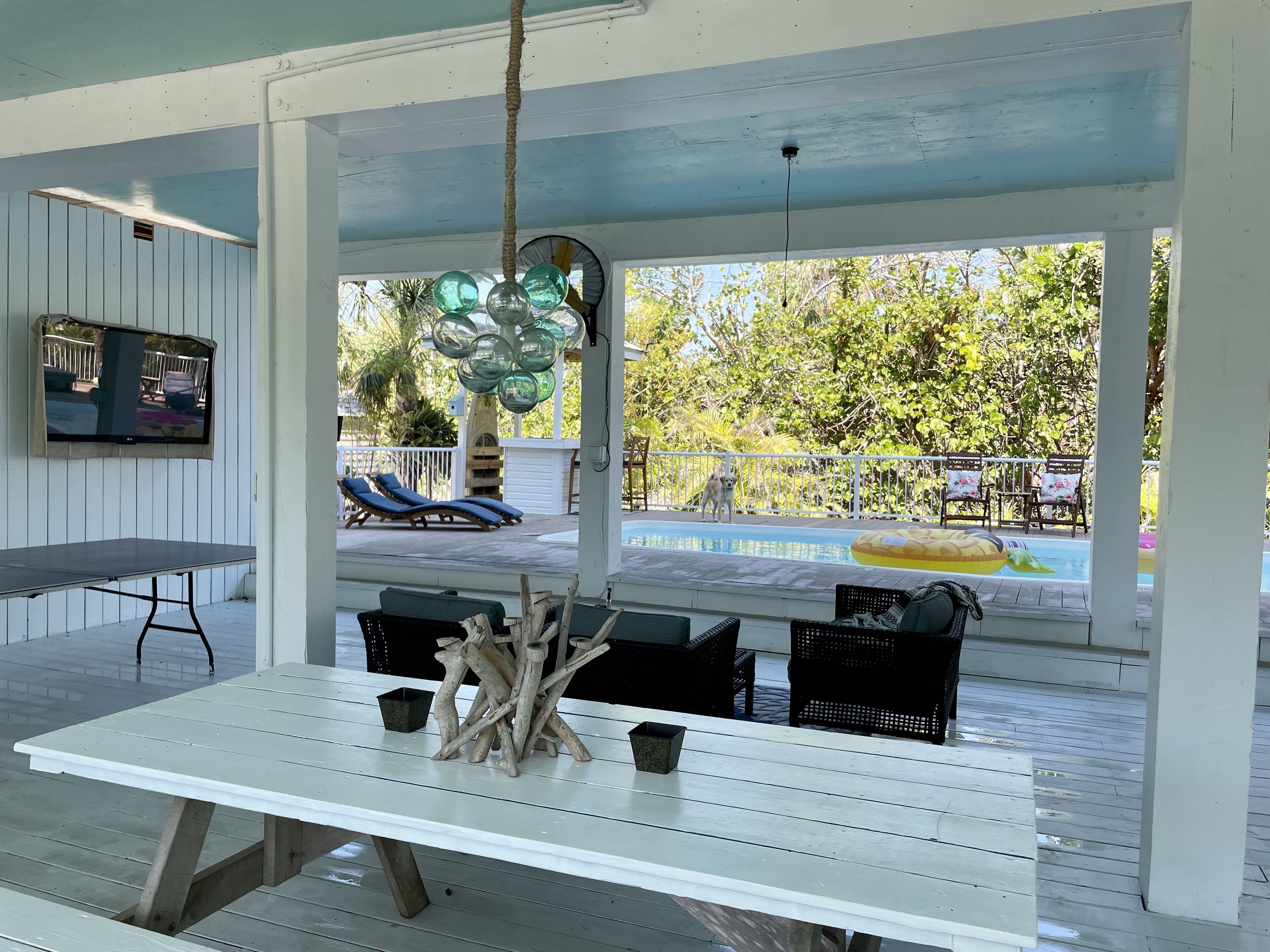 A covered outdoor area with a pool, lounge chairs, a table, and a TV screen.