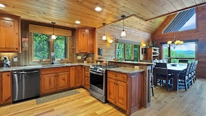 A spacious kitchen with wooden cabinets, a stainless steel dishwasher, and a large island with a stove. There is a dining area with a table and chairs, and large windows offering views of the outdoors. - Alpine Ridge - Mountain Views | Close to Down Town Blue Ridge | Hot Tub | Shuffleboard (Blue Ridge)