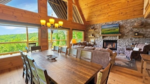 A wooden interior with a dining table, chairs, a stone fireplace, and a view of greenery outside. - Alpine Ridge - Mountain Views | Close to Down Town Blue Ridge | Hot Tub | Shuffleboard (Blue Ridge)