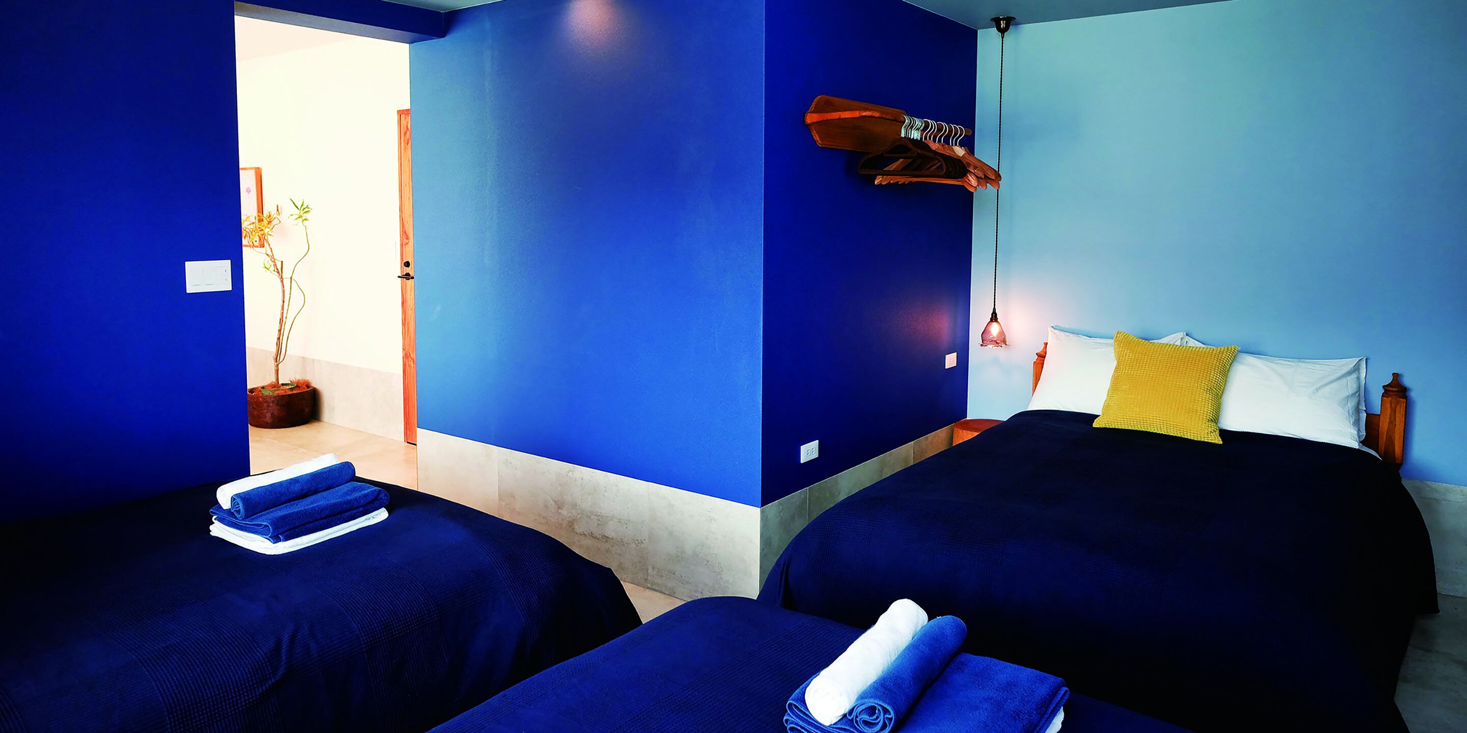 Une chambre d’hôtel avec deux lits, une tête de lit en bois, un luminaire suspendu et une plante en pot.