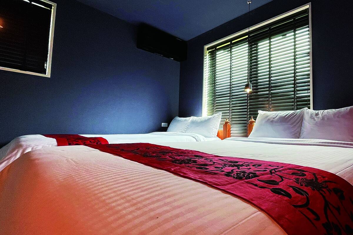 Quarto de hotel com uma cama grande, uma janela com persianas, uma mesa de cabeceira com abajur e um ar condicionado fixado na parede.