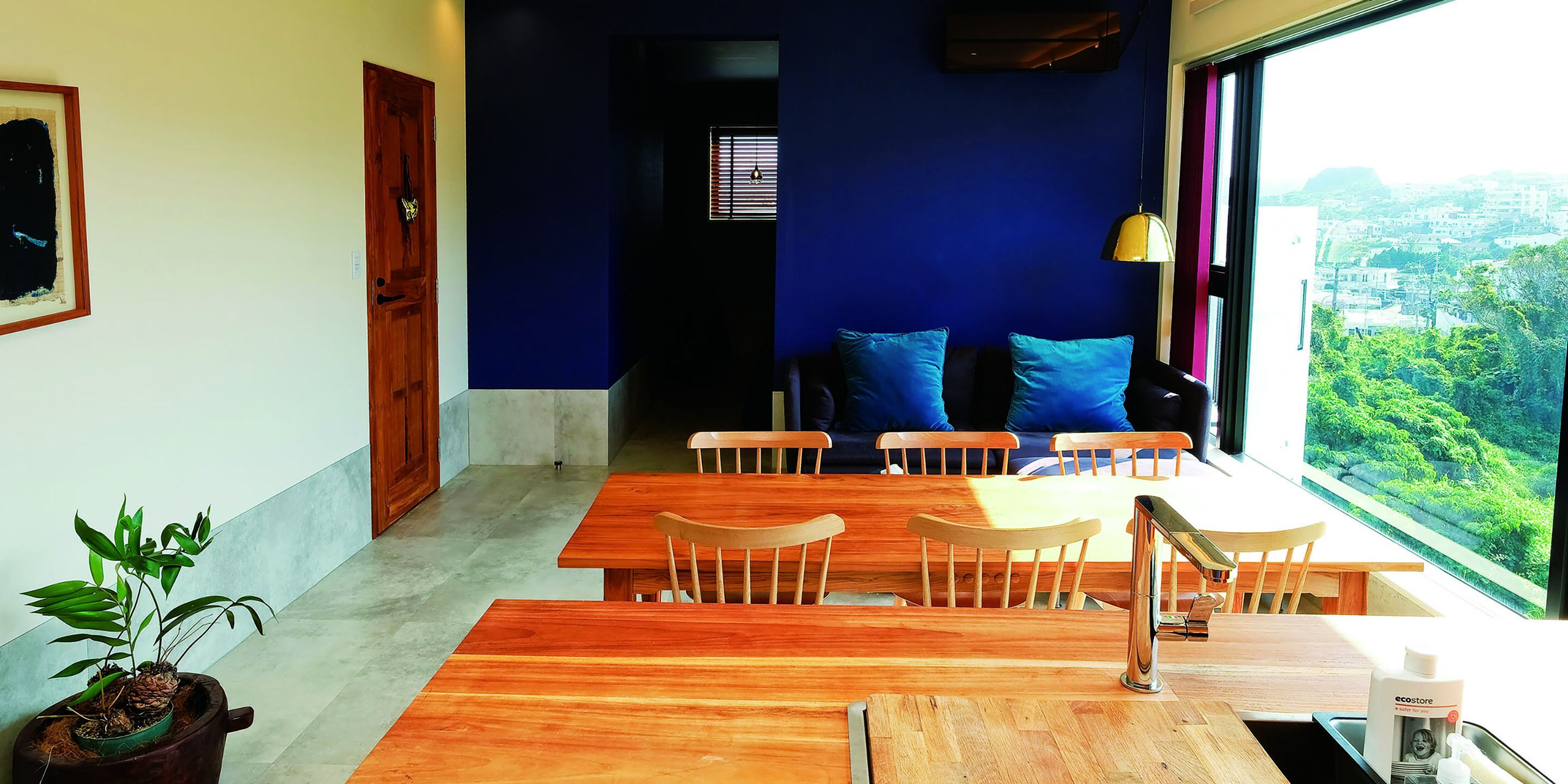 Un comedor con mesas y sillas de madera, una planta en maceta y vistas al verdor por la ventana.