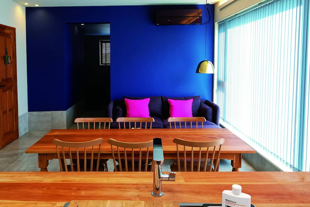 Uma área de refeições com uma mesa e cadeiras de madeira, uma luminária pendente moderna e um sofá com almofadas coloridas.