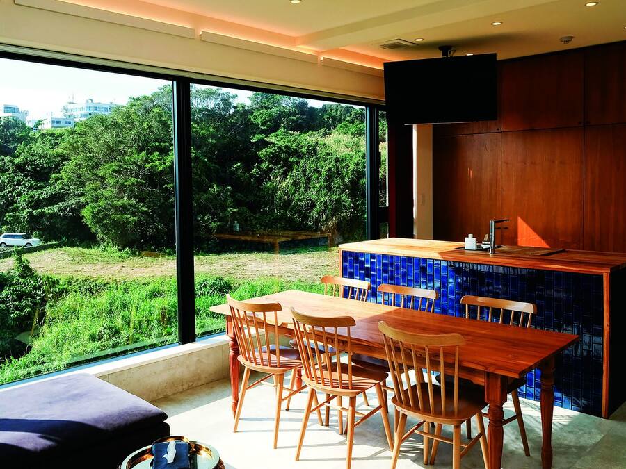 A modern kitchen with a wooden table and chairs, a blue-tiled backsplash, and a large window overlooking a green landscape.