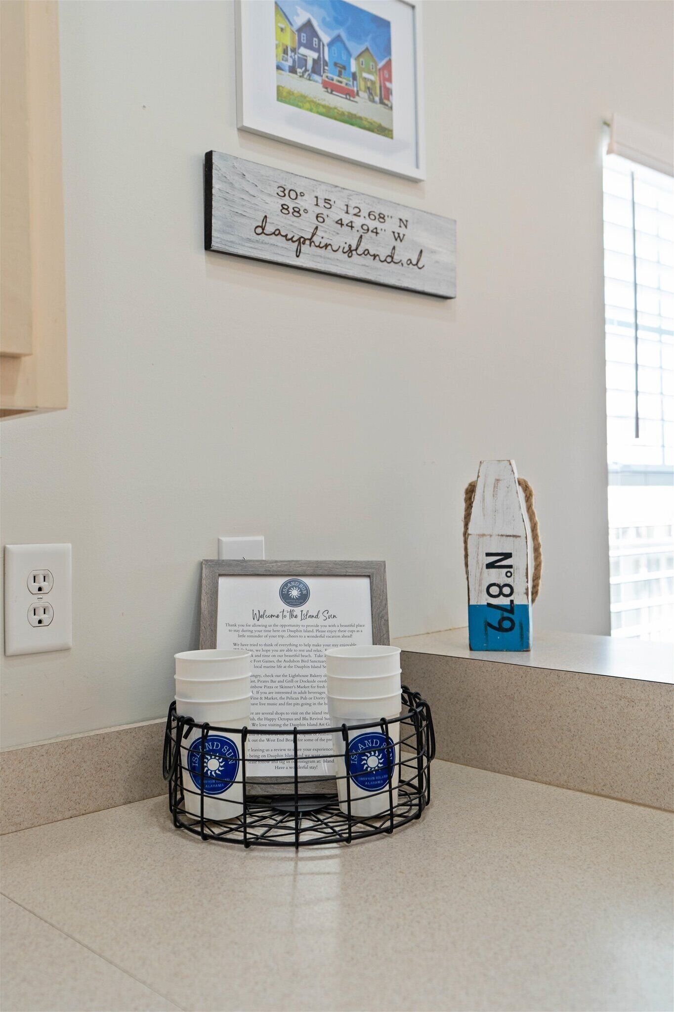 A kitchen counter with a framed welcome sign, a wooden sign with coordinates, and a decorative bottle.